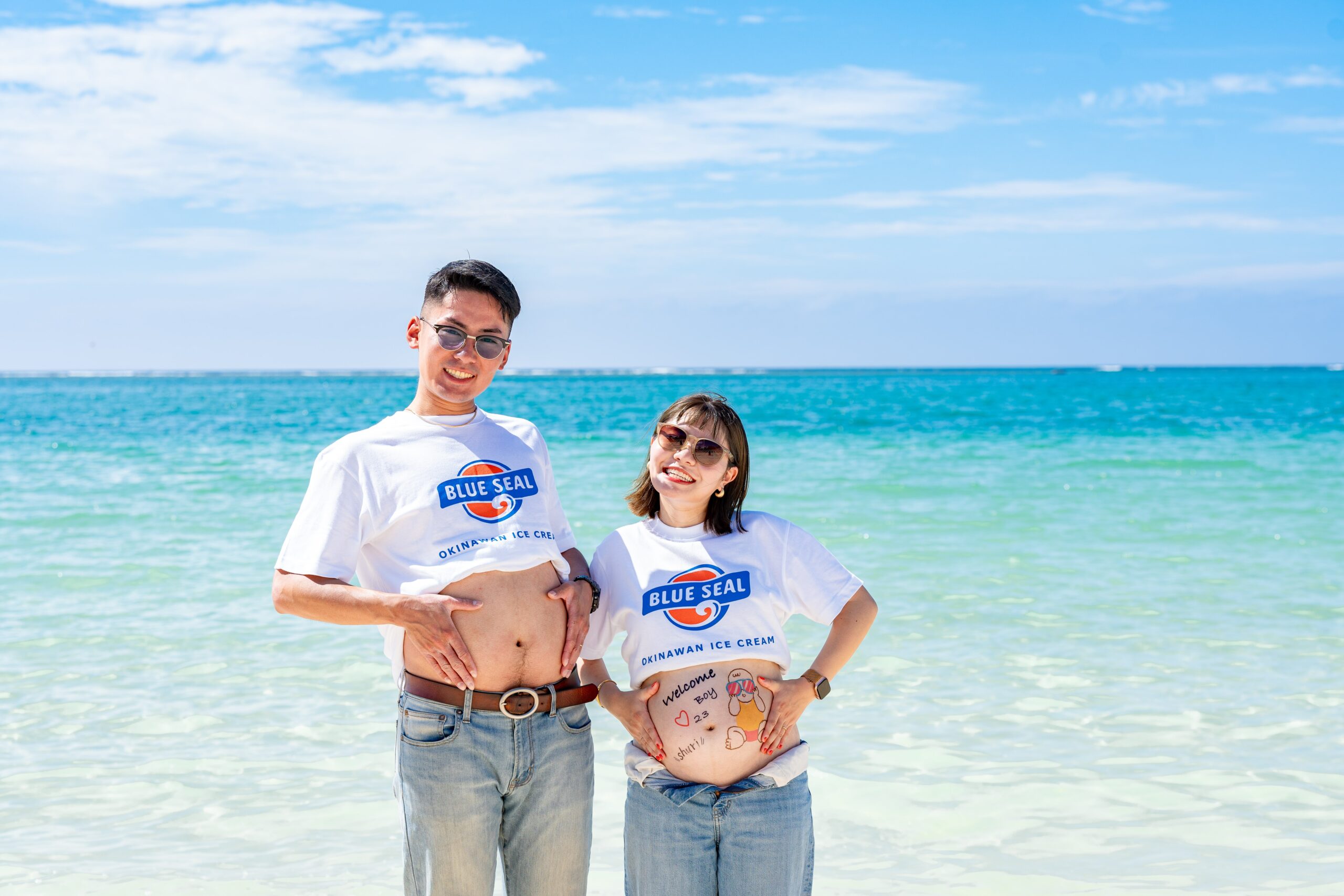 沖縄のビーチで、お揃いのブルーシールTシャツを着てお腹出しポーズを楽しむ夫婦のマタニティフォト。