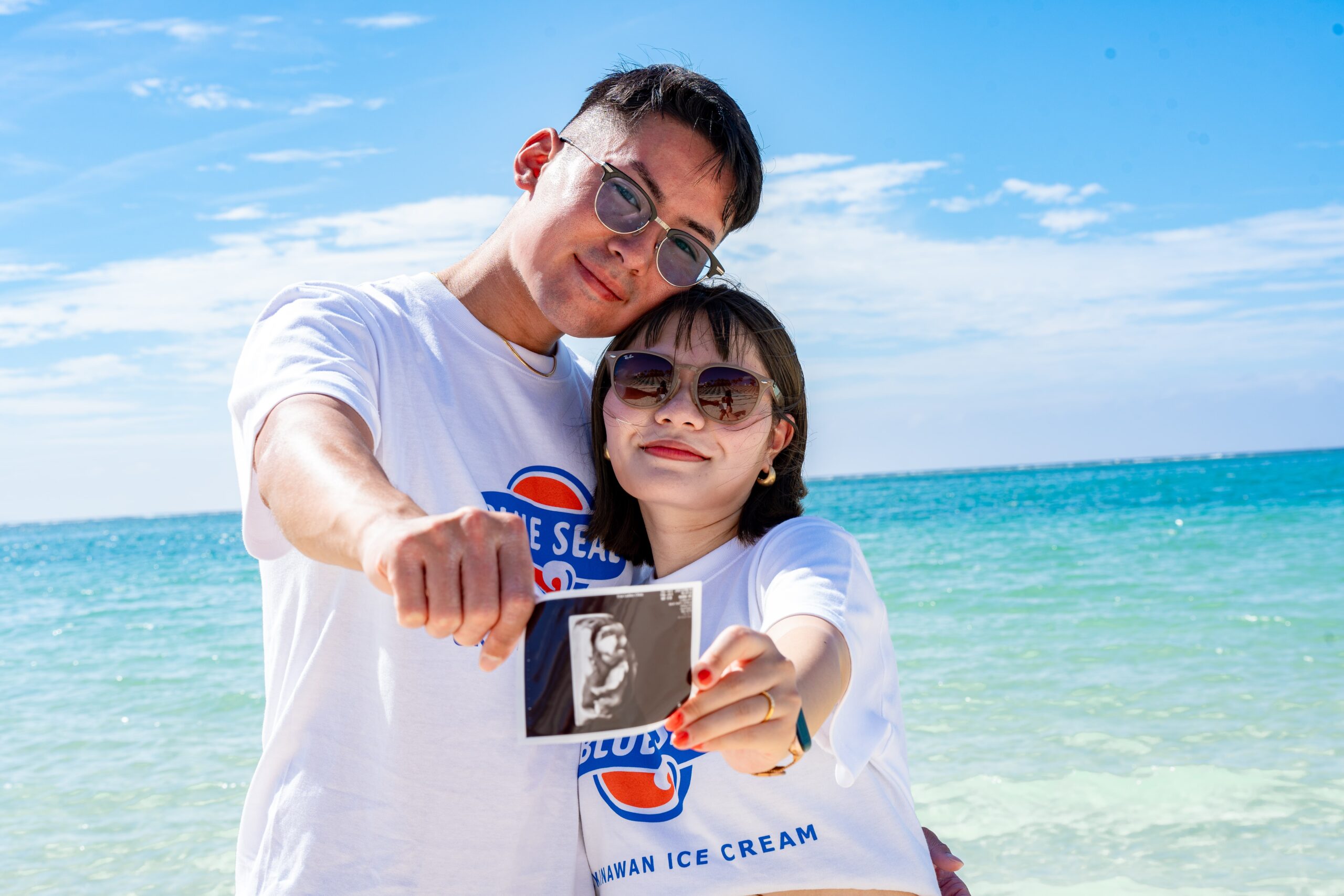 沖縄の海辺で、お揃いのTシャツを着た夫婦がエコー写真をカメラに突き出して喜ぶマタニティフォト。