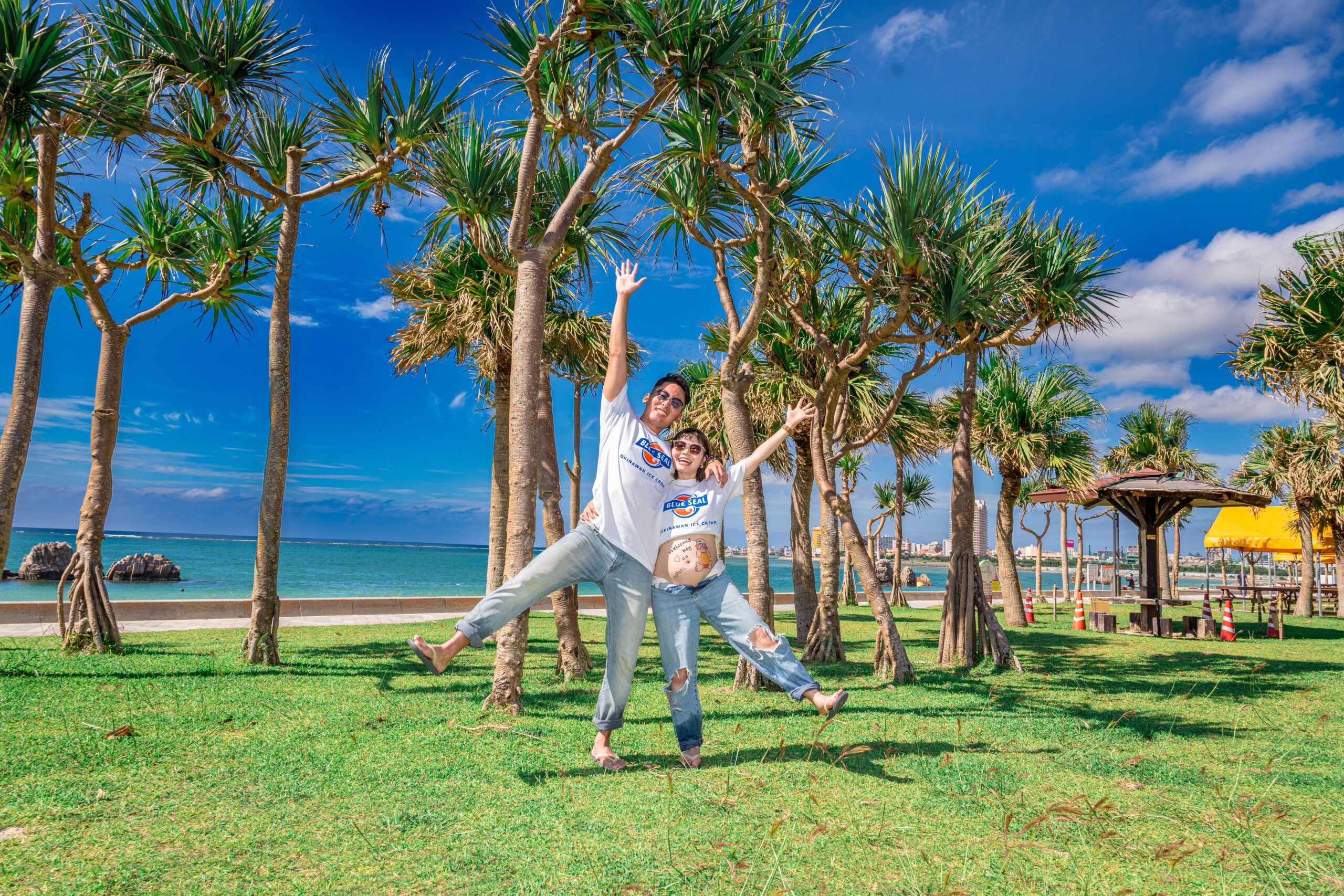 沖縄のヤシの木がある芝生エリアで、夫婦が元気にジャンプする躍動感あふれるマタニティフォト。