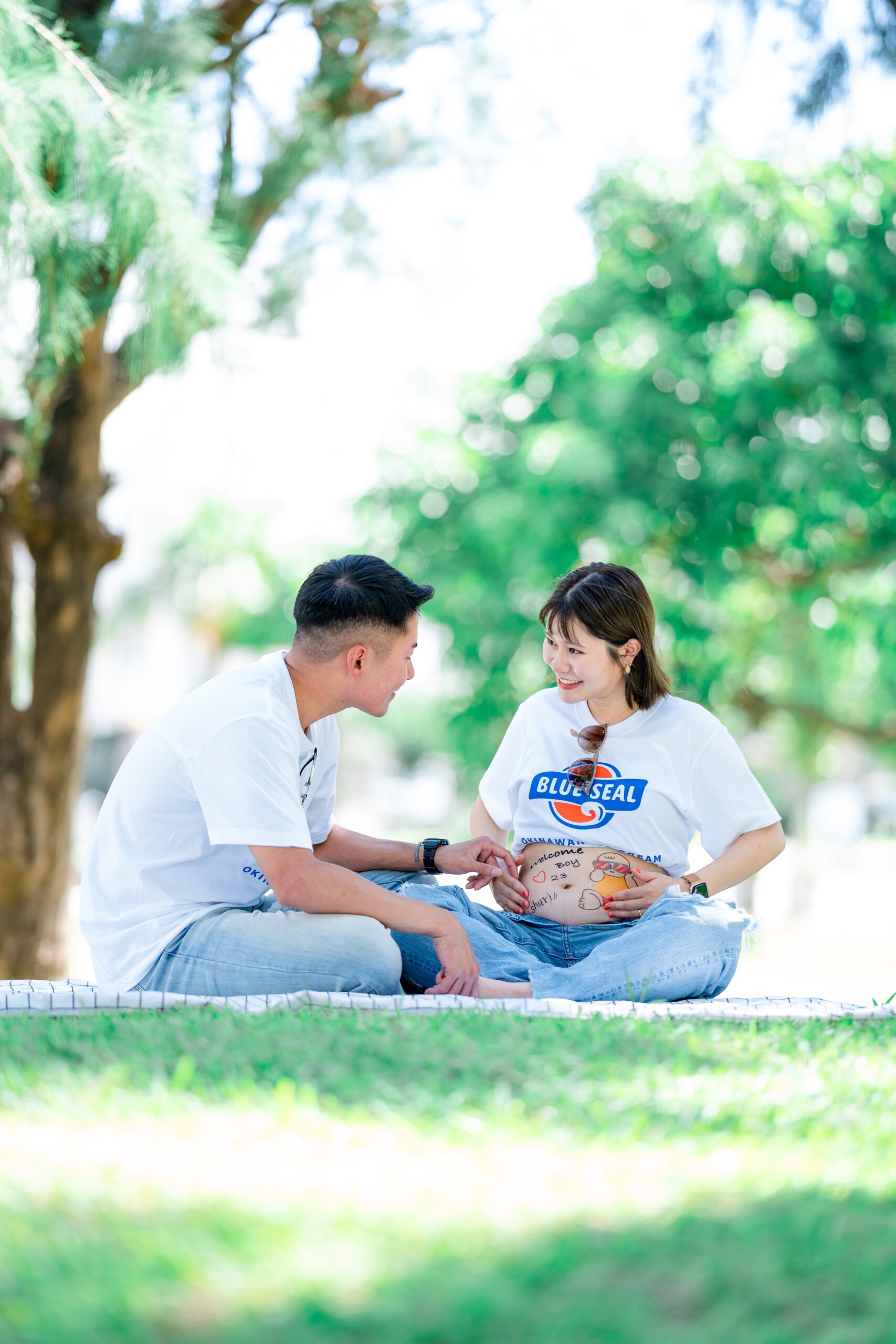 沖縄の木陰にレジャーシートを敷き、夫婦で座りながらお腹を見つめ合う自然体なマタニティフォト。