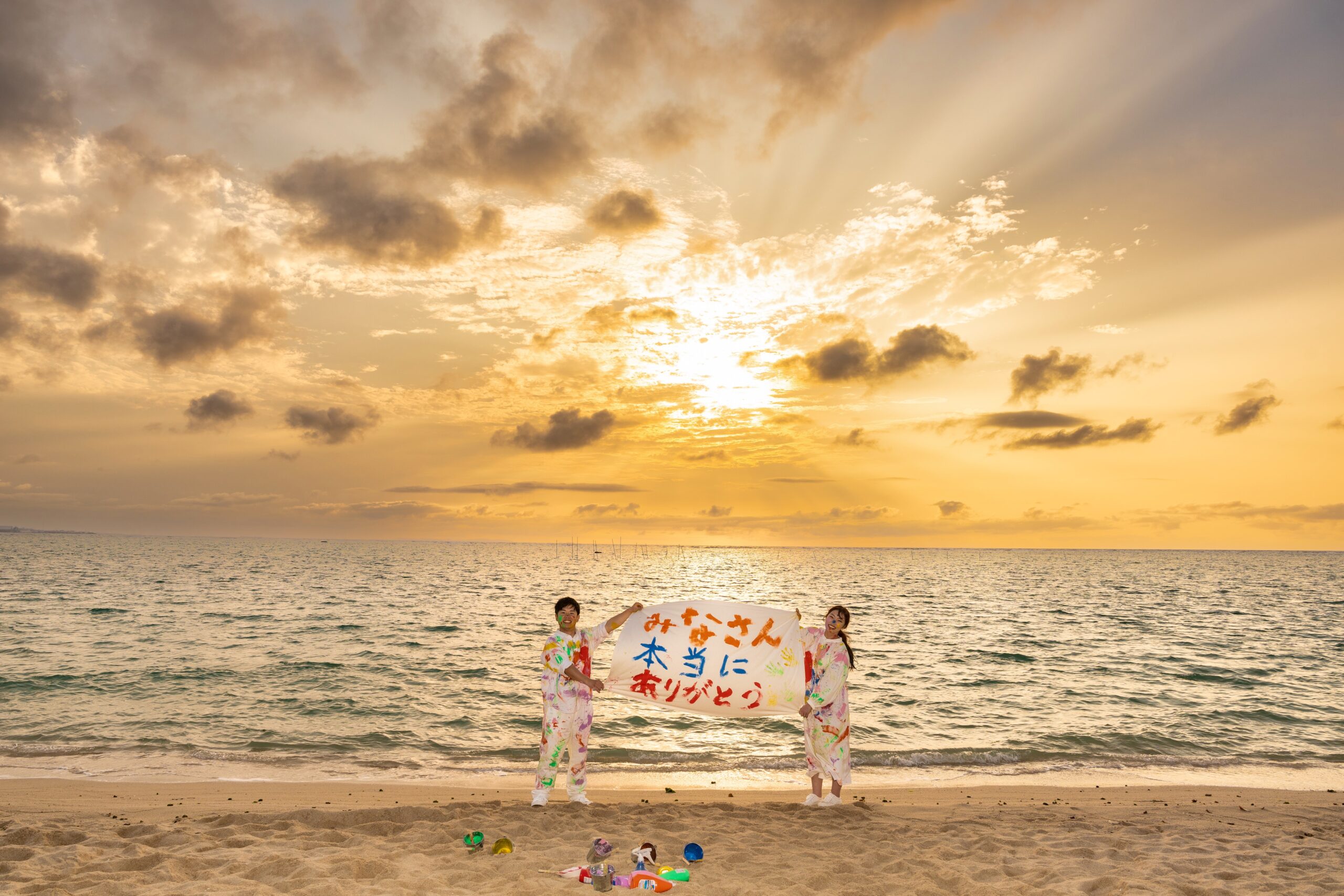 沖縄ペイントフォト｜結婚式用の横断幕を掲げるカップルのアラハビーチ前撮り撮影