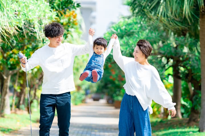 沖縄の公園でパパとママに腕を持ち上げられジャンプする子どもの家族写真｜沖縄家族写真