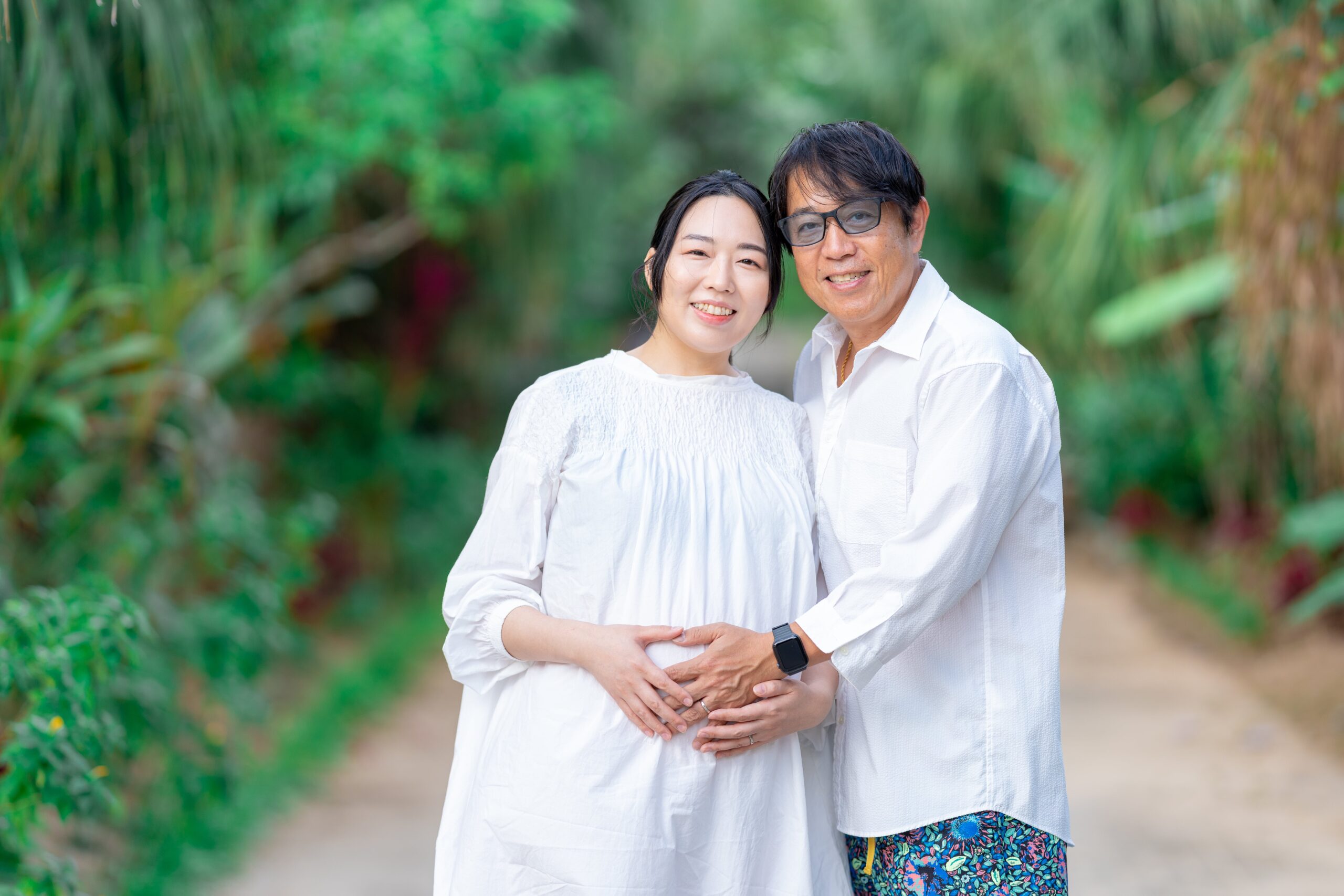 沖縄の木漏れ日が差し込む並木道で、夫婦が笑顔で見つめ合うナチュラルなマタニティフォトのワンシーン。