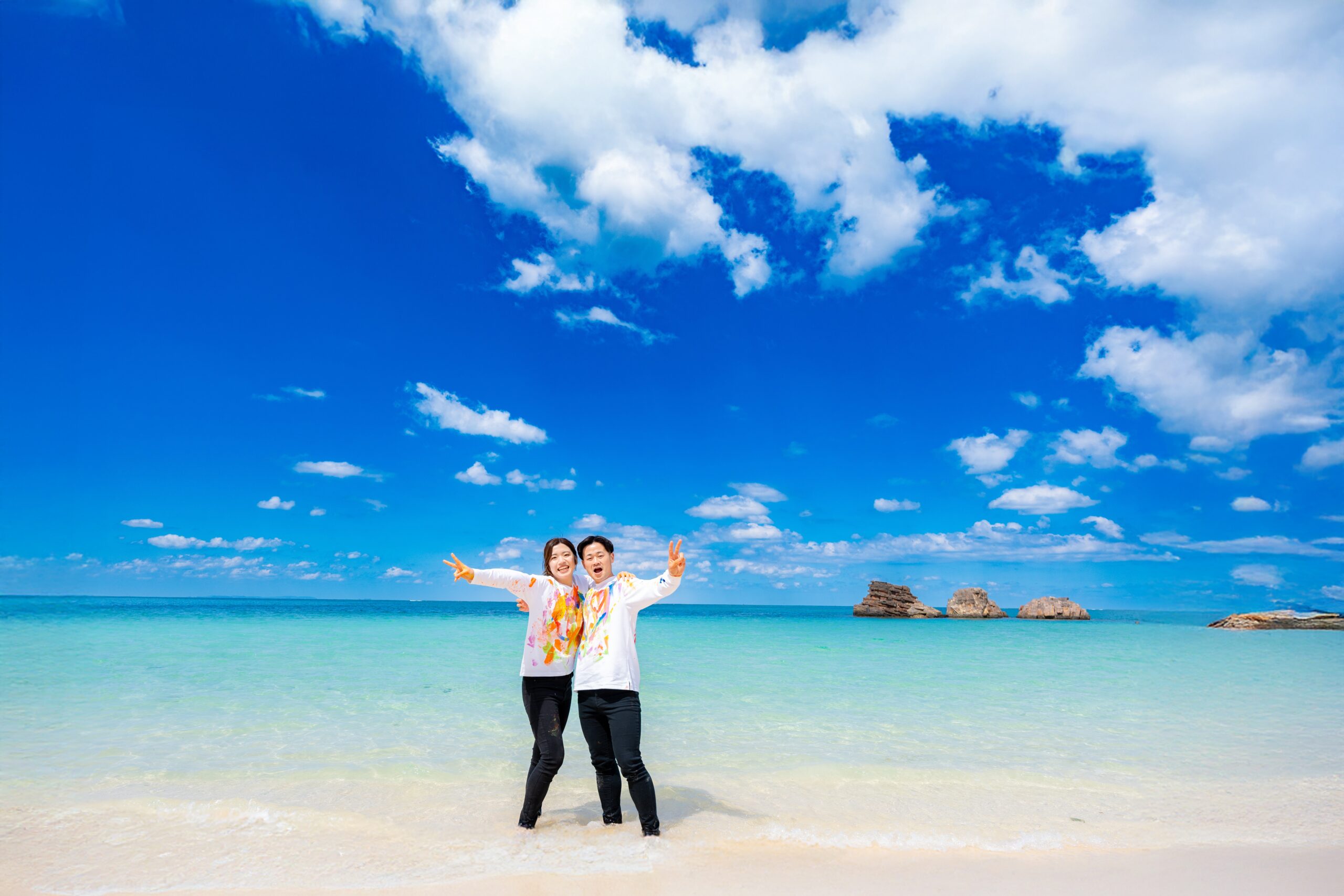 沖縄ペイントフォト｜青い海でピースサインの絵の具まみれカップルのビーチフォト