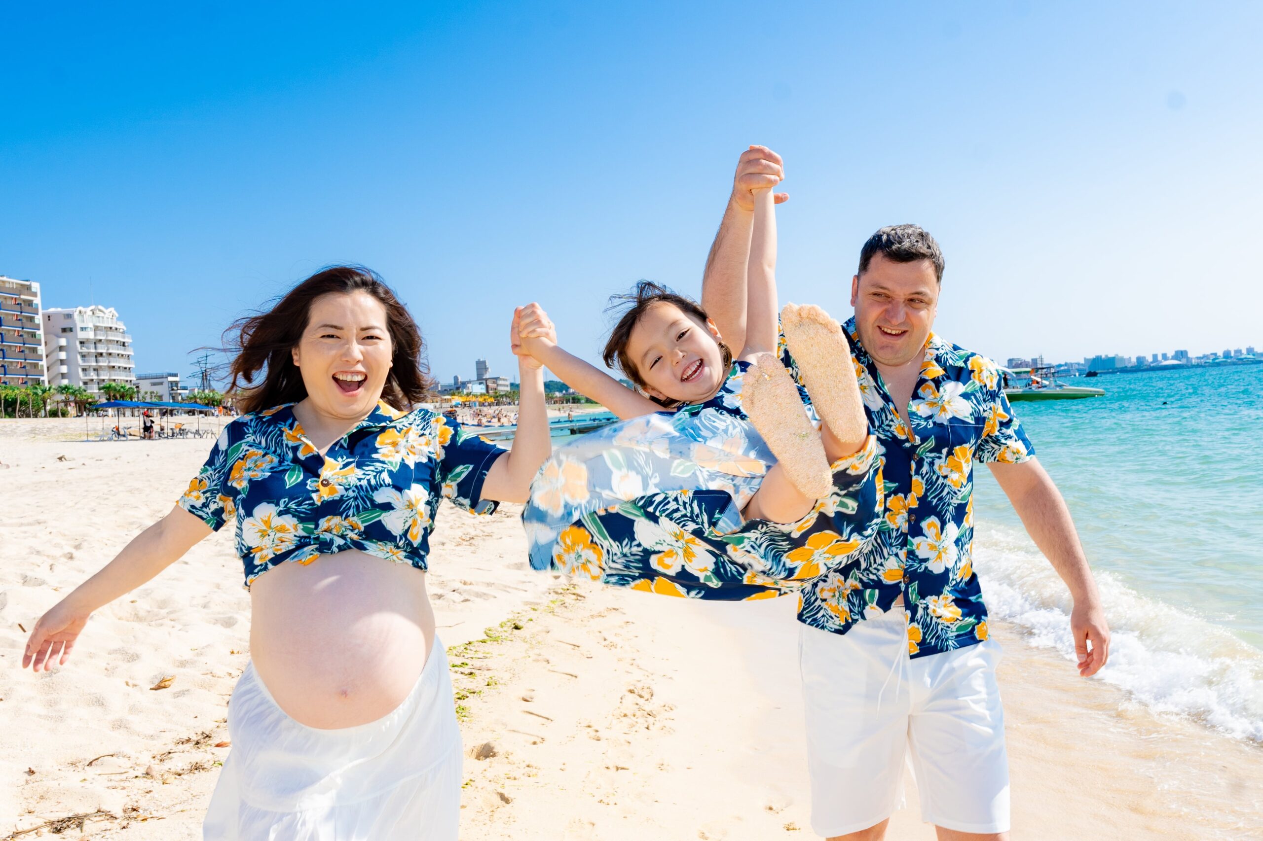 沖縄のビーチで、お揃いのかりゆしウェアを着た家族3人が海辺で上の子を高く持ち上げて笑い合う、躍動感のあるマタニティフォト。
